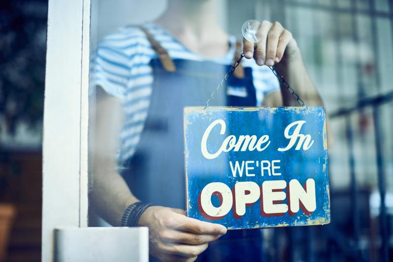 一个穿着工作服的人在商店橱窗里举着一块蓝色牌子，上面写着：“Come in, we're open（请进，我们营业中）”
